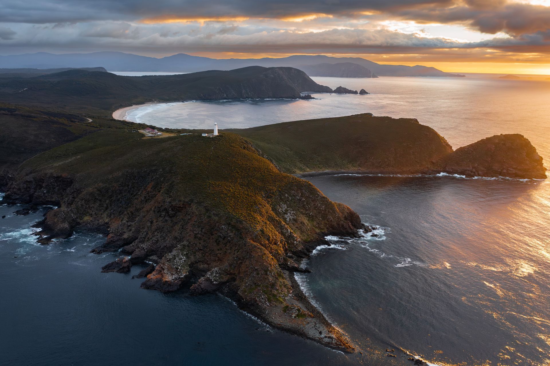Aerial view of a rugged coastline with dark green hills, a bay, and an ocean at sunset.