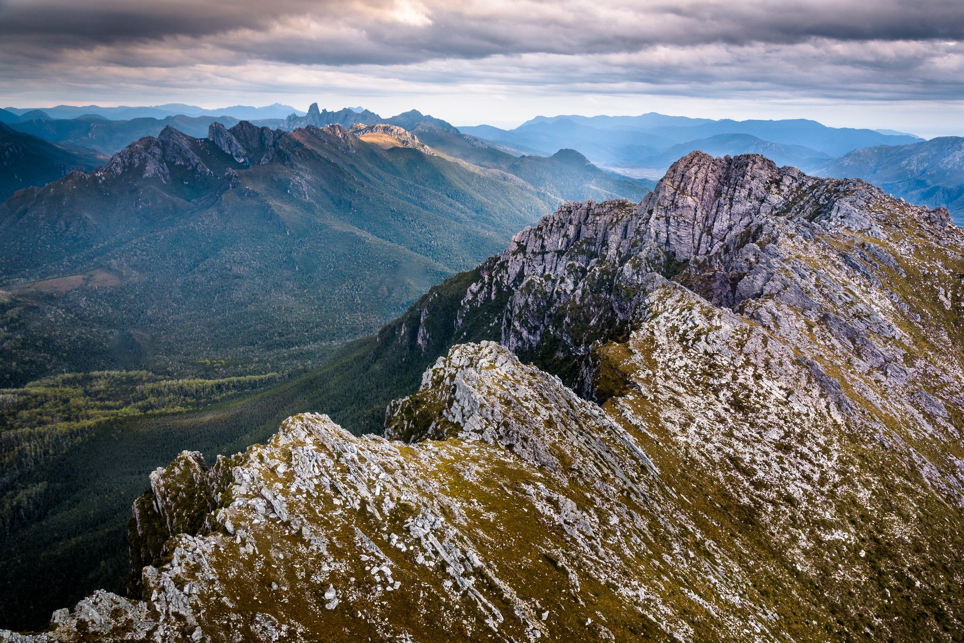Rugged mountain range with rocky peaks and green forested valleys under a cloudy sky.