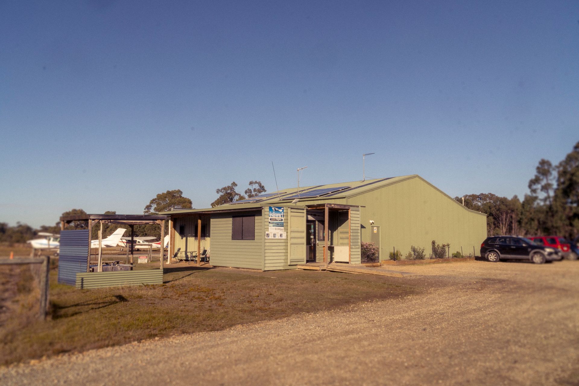 A small green building with a porch and attached metal structure, parked cars in the background, a clear blue sky.