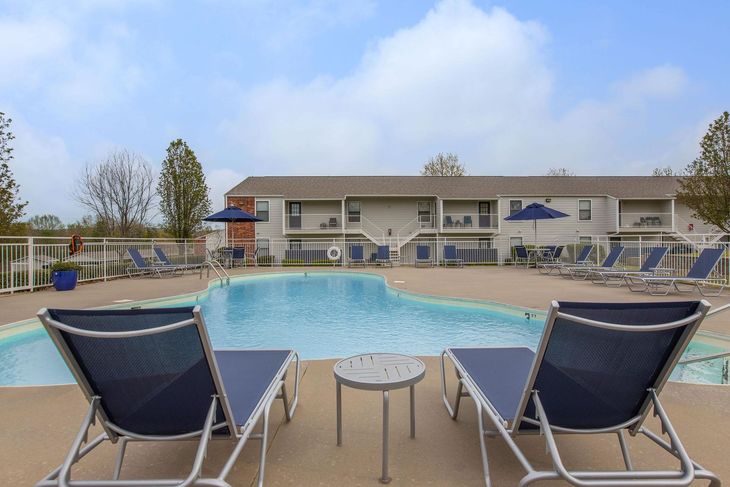Poolside lounge chairs facing a pool, with a building in the background under a cloudy sky.