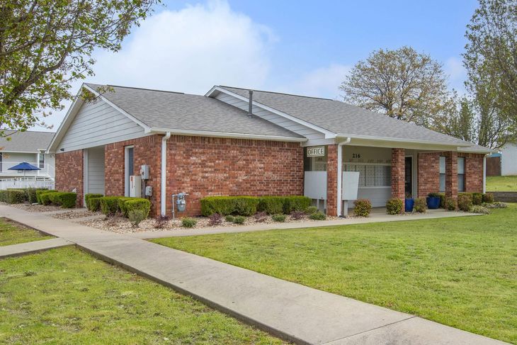 Brick building with grey roof, bushes along the front, and a sidewalk on a grassy lawn.