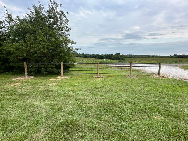 Grassy field with a wooden fence, trees on the left, and water under a cloudy sky