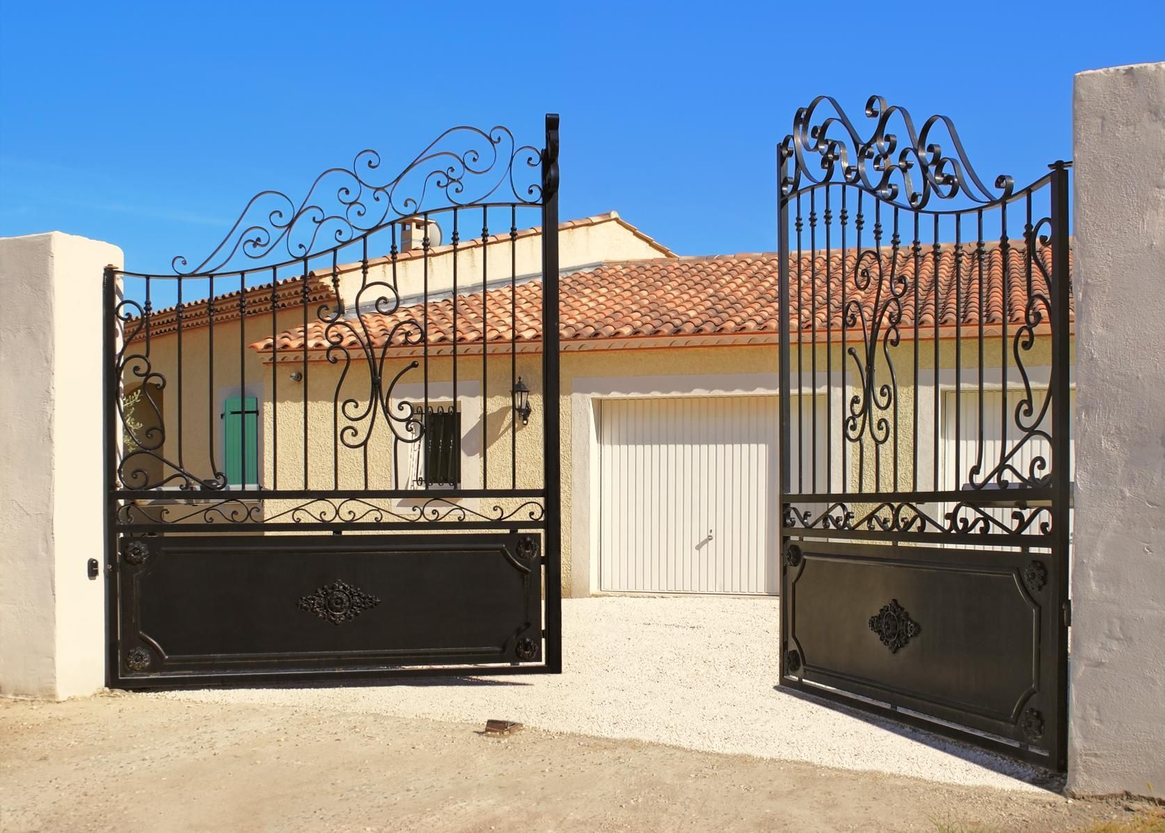 Open wrought-iron gate leading to a sunlit house with a tiled roof and blue sky
