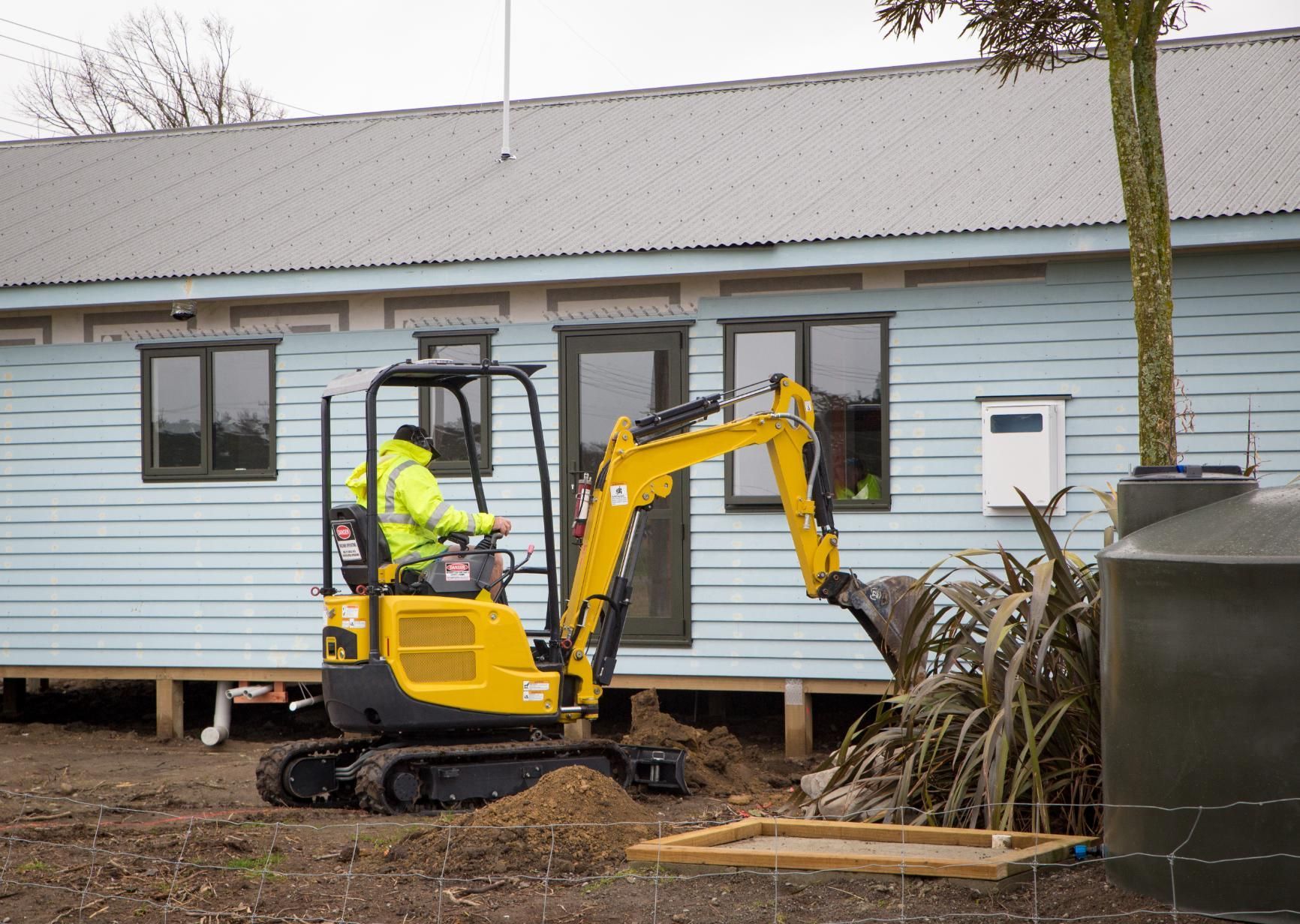 Yellow mini excavator and worker demolishing a blue house wall outdoors