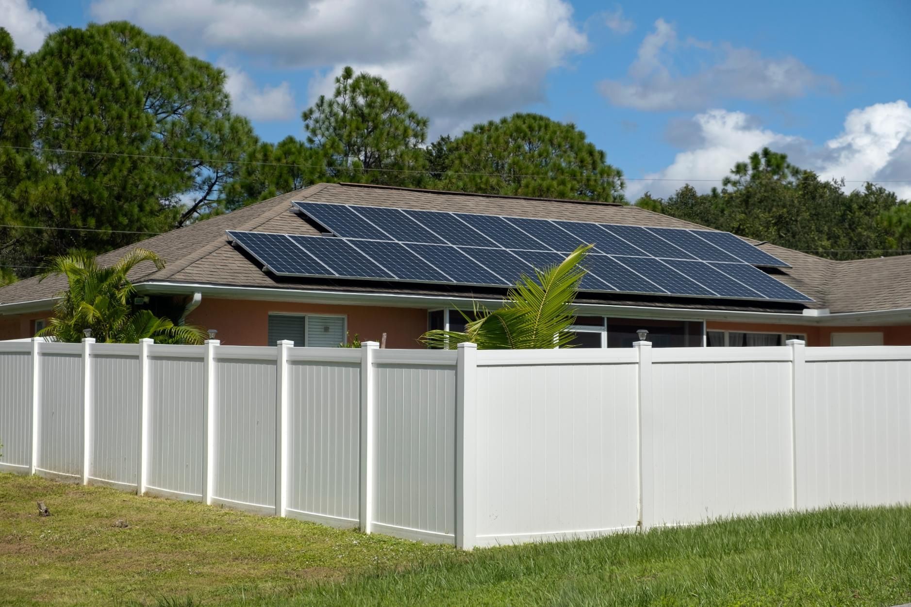 Suburban house with rooftop solar panels behind a white fence on a sunny day