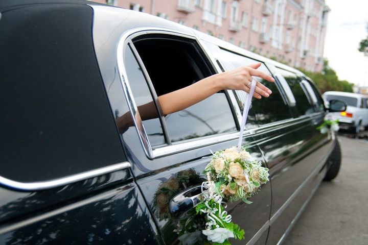 A person reaches from a black limousine, holding a bouquet. The setting appears to be outdoors near a building.
