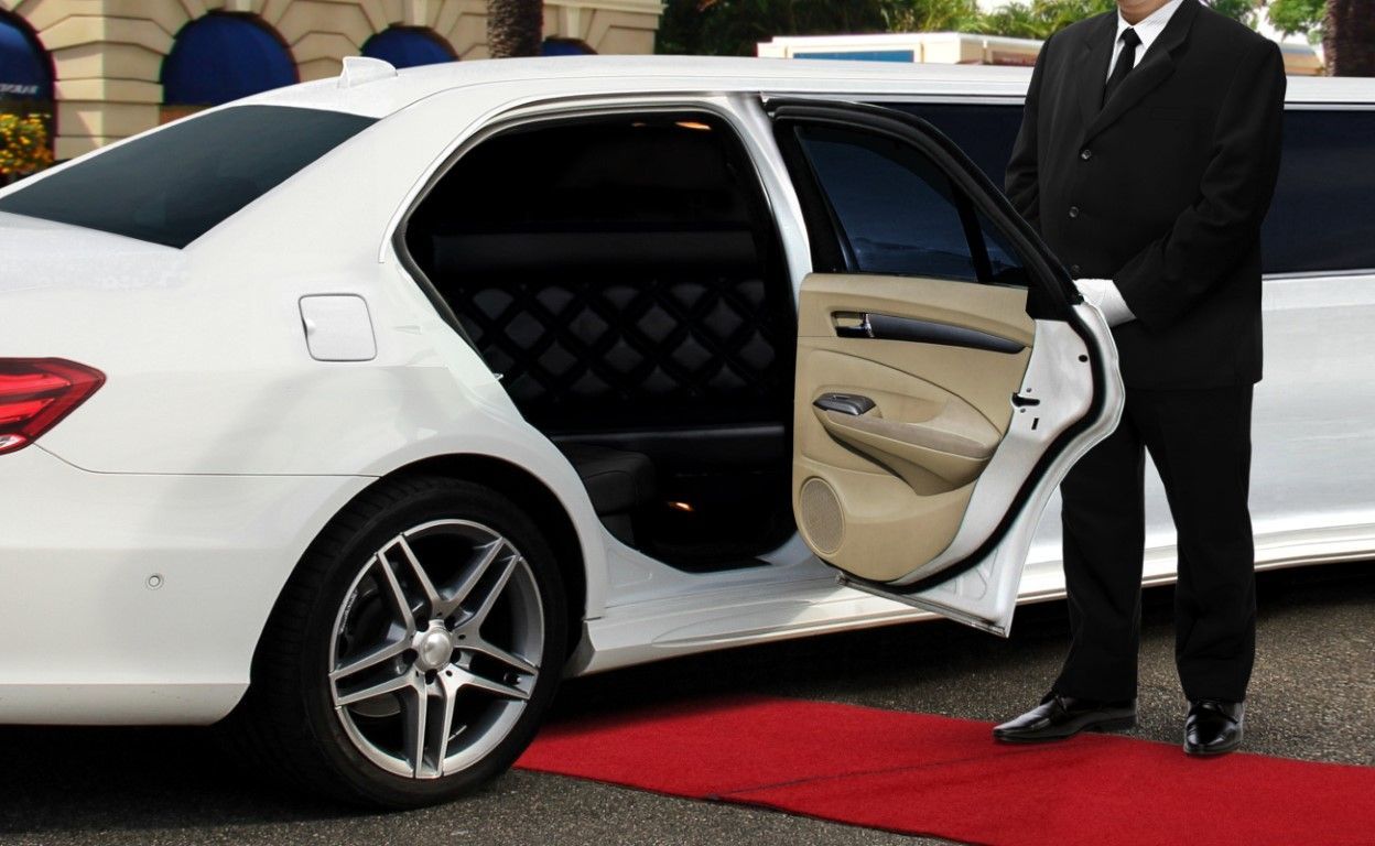 A chauffeur in a black suit opens the door of a white limousine onto a red carpet.