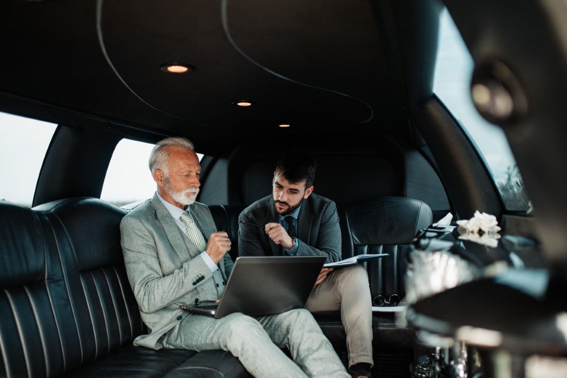 Two men in suits inside a black limousine, one using a laptop, the other holding a notepad, discussing business.