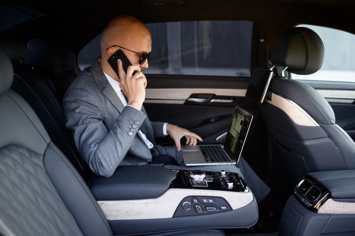 A businessman in a car's backseat, wearing a suit and sunglasses, uses a laptop and phone. The car interior is dark with light leather seats.