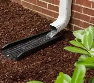 Downspout draining water into a black, grooved ground drain in brown mulch next to a brick wall.