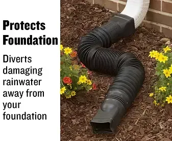 Black corrugated drainpipe extending from a white gutter downspout, diverting water away from a brick foundation and flowerbed.