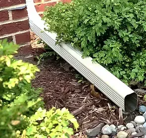 White gutter emptying water into a landscaped area with mulch, rocks, and green bushes. Brick wall in background.