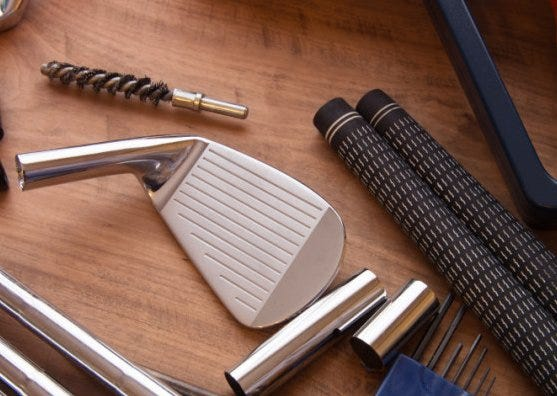 Golf club iron head and 2 golf grips on a club repair workbench. Club repair tools are also nearby on the workbench.