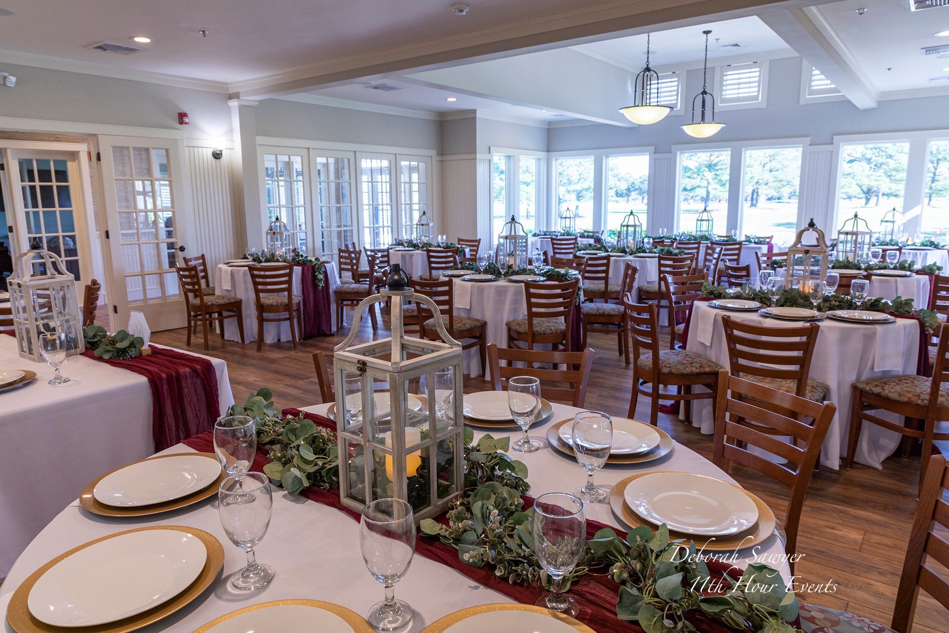 Formal dining room set for a wedding reception with round tables, white linens, and burgundy accents. Wooden chairs surround the tables, each decorated with greenery and lanterns.