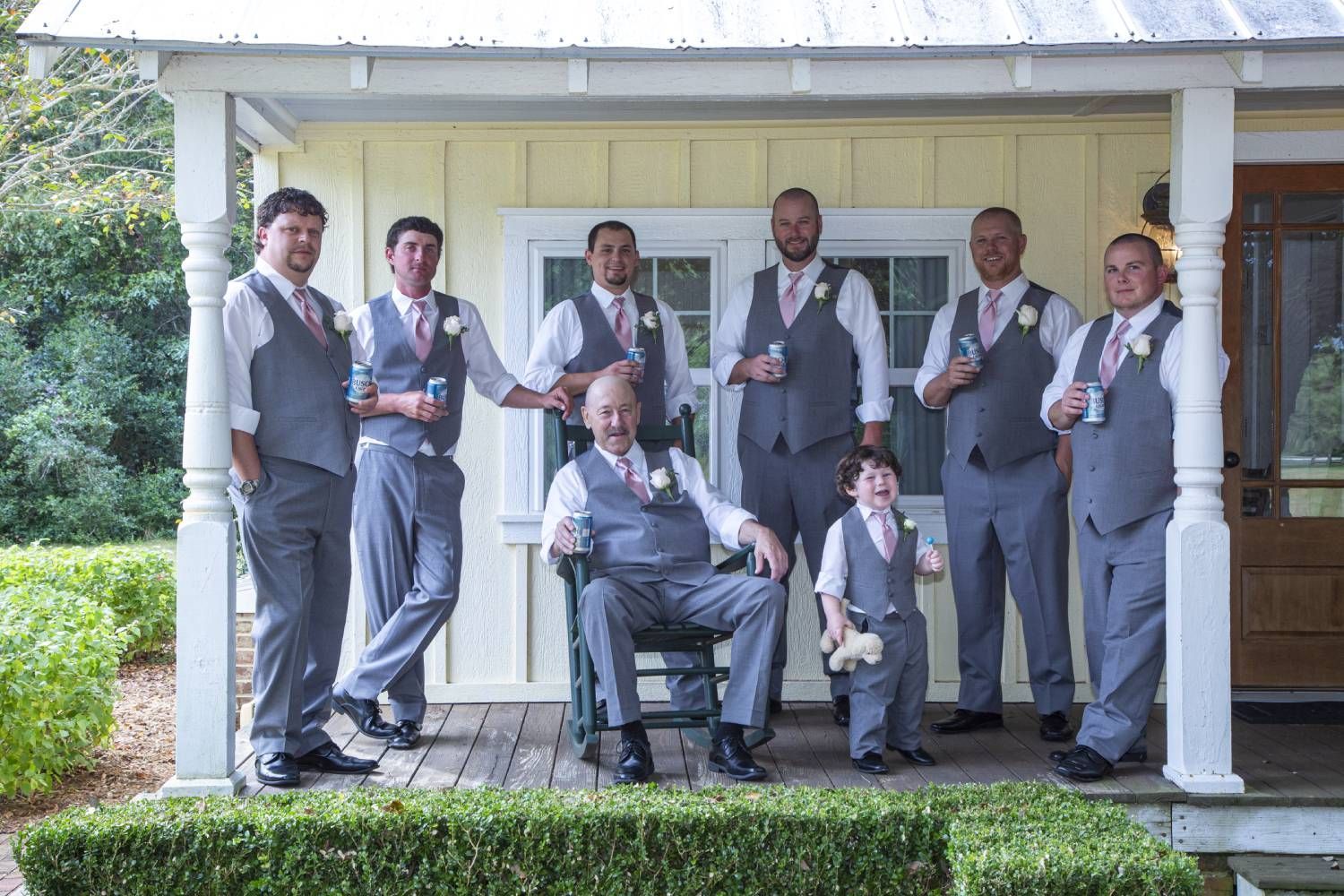 A group of men, including a child, in gray suits pose on a porch in front of a yellow building. They are smiling and holding drinks.