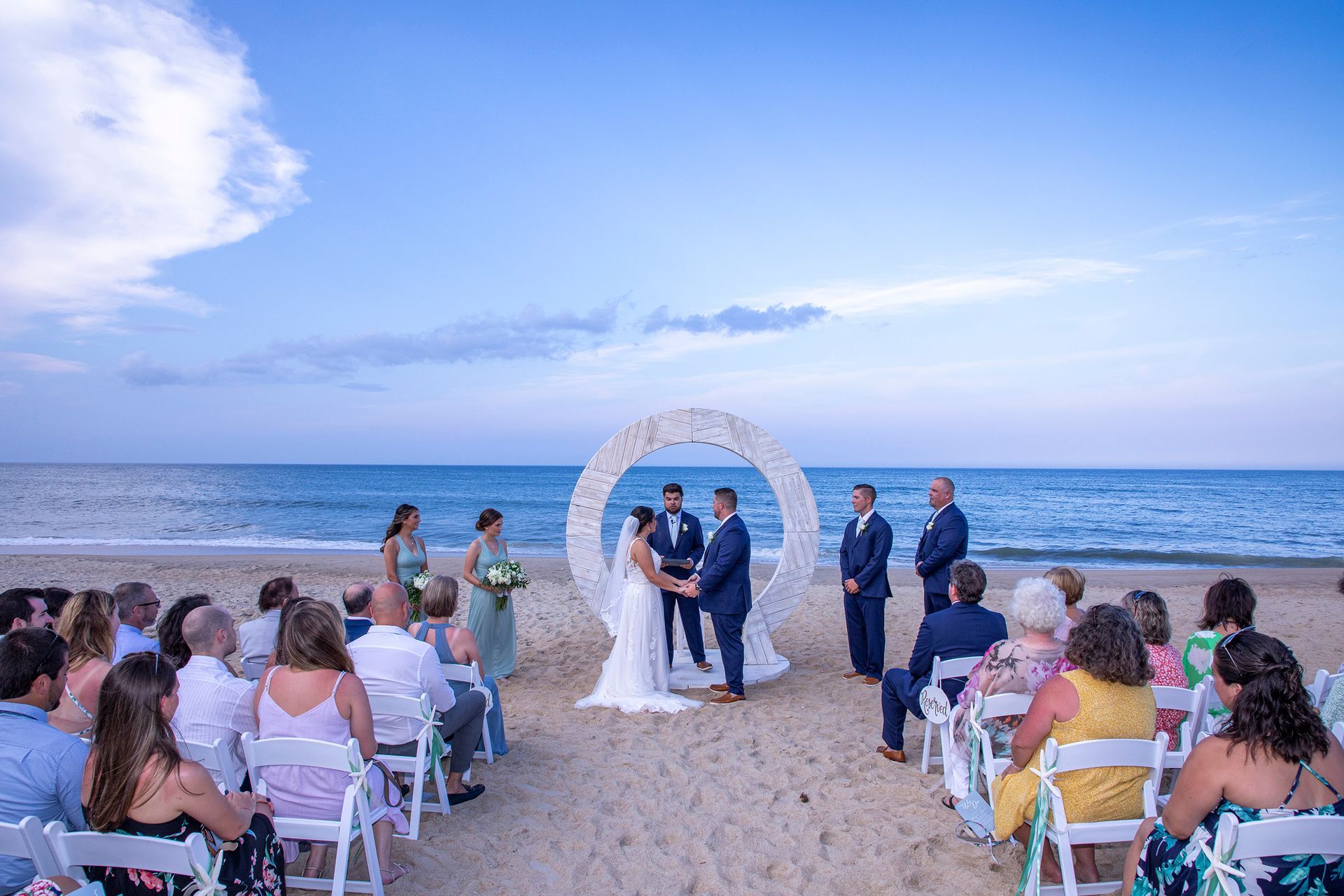 A wedding ceremony on a beach at dusk. A couple holds hands under a floral arch, surrounded by guests in chairs.