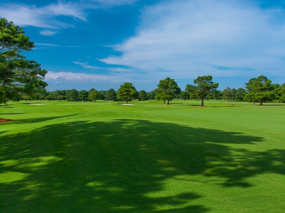 Green golf course under a blue sky with scattered trees and shadows.