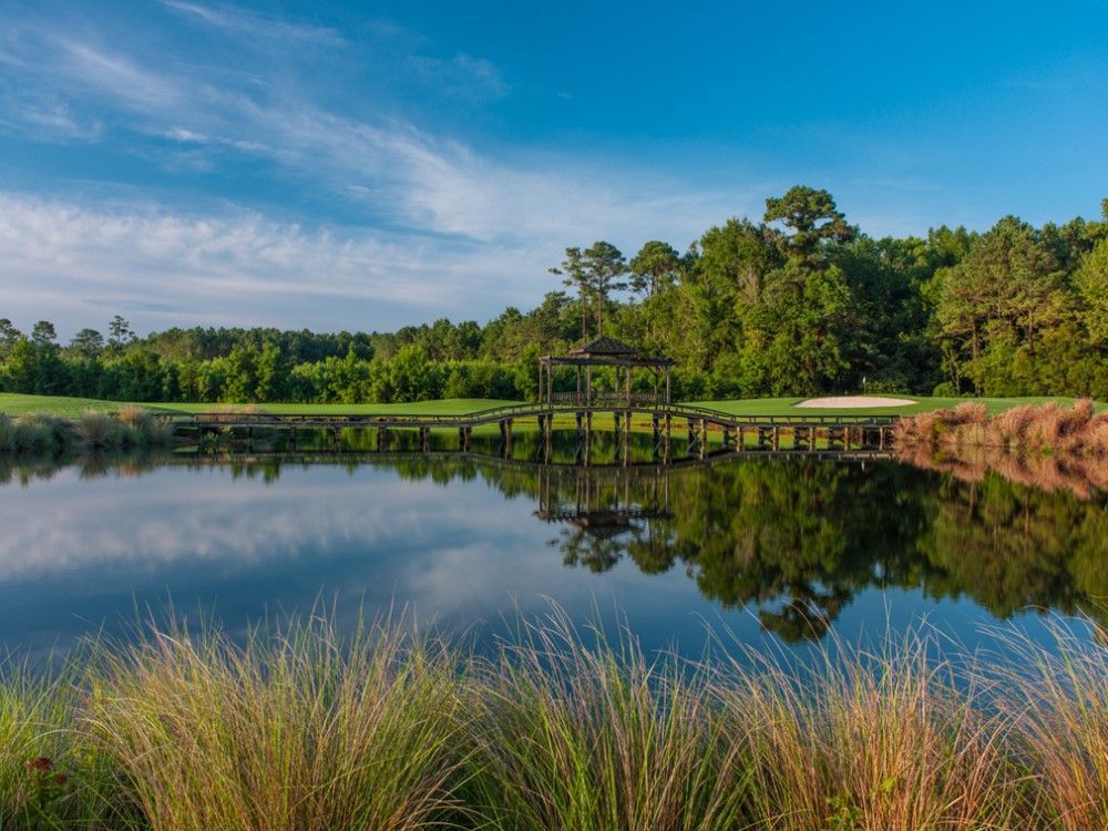 A serene landscape of a golf course with a reflective lake, a gazebo, and lush greenery under a bright blue sky.
