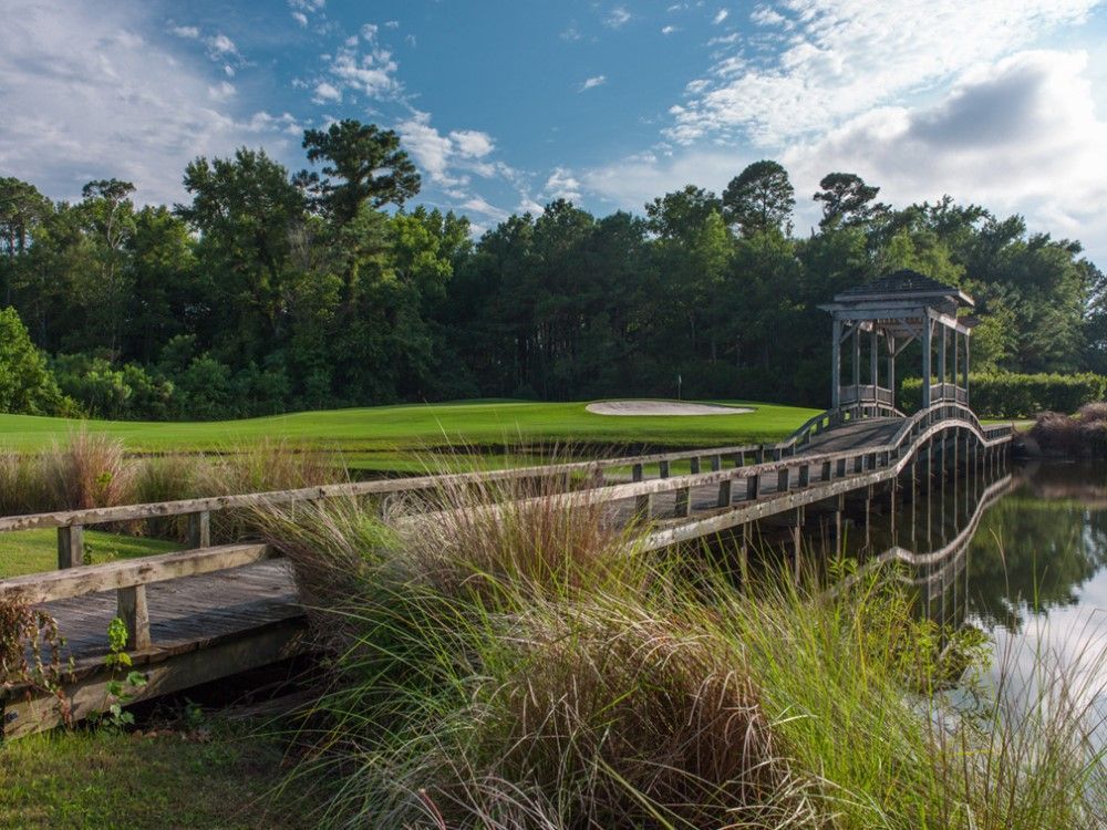 Wooden bridge with a gazebo over water, leading to a golf course on a sunny day.