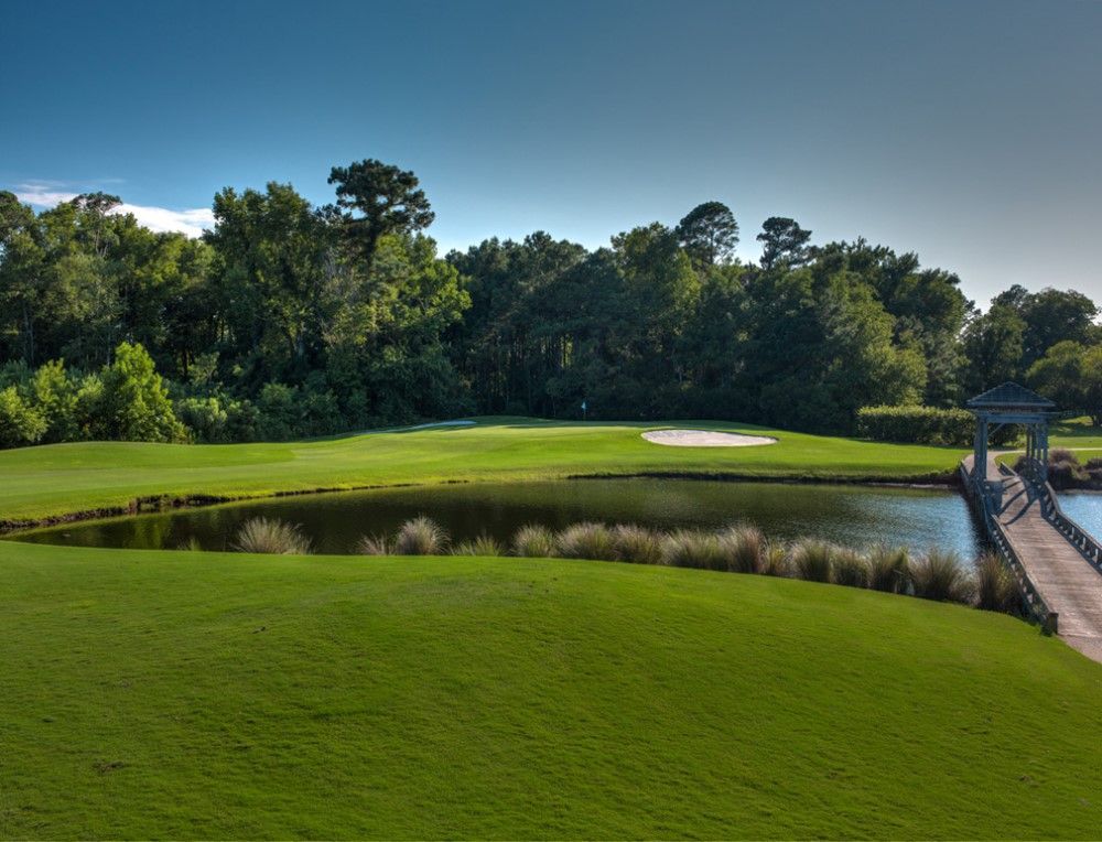 Green golf course with a pond in the foreground, a sand trap, and a gazebo. Trees and a blue sky are in the background.