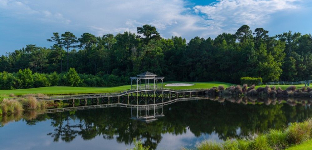 A gazebo sits on a bridge over a calm lake, surrounded by green grass and trees. The sky is partly cloudy.