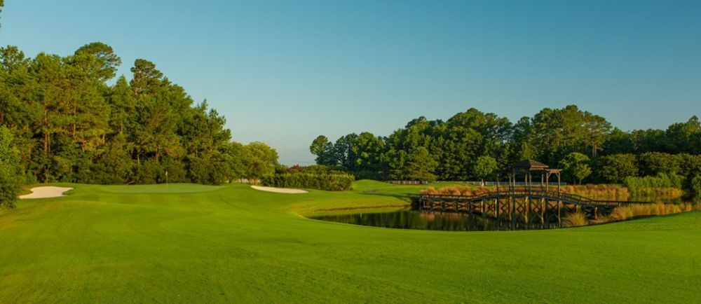Green golf course with a pond, bridge, and trees under a blue sky.