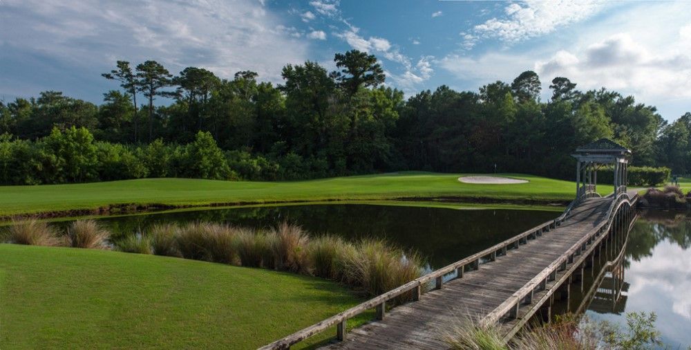A wooden bridge over a pond leads to a golf green, with trees in the background under a cloudy blue sky.