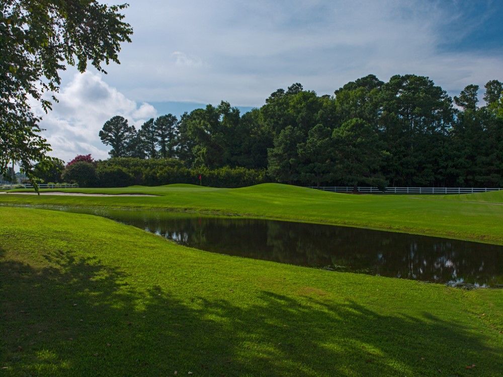 Grassy golf course with a pond, trees, and a partly cloudy sky. Sunlight casts shadows on the lawn.