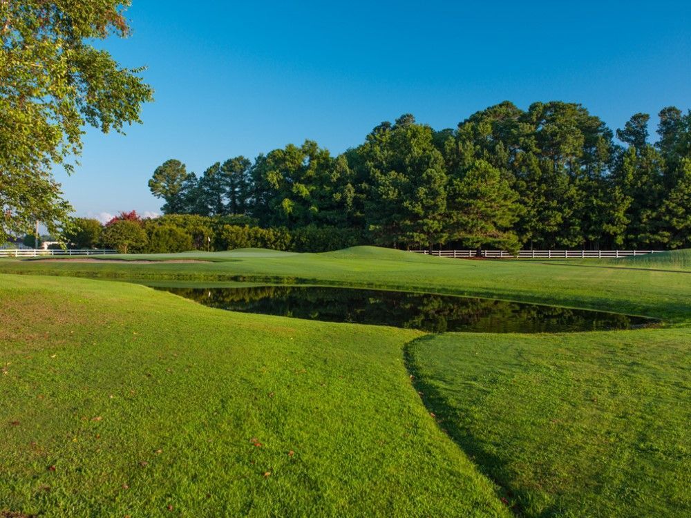 Green golf course with a pond and trees under a clear blue sky.