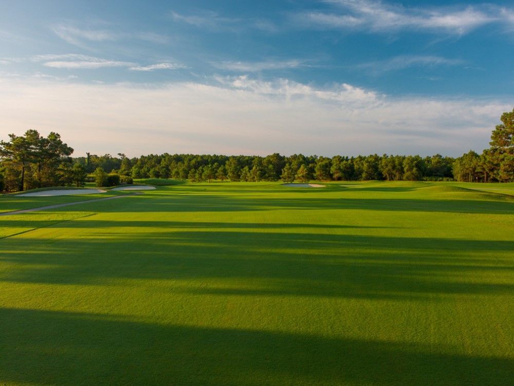 A green golf course bathed in sunlight with a backdrop of trees under a partly cloudy blue sky.