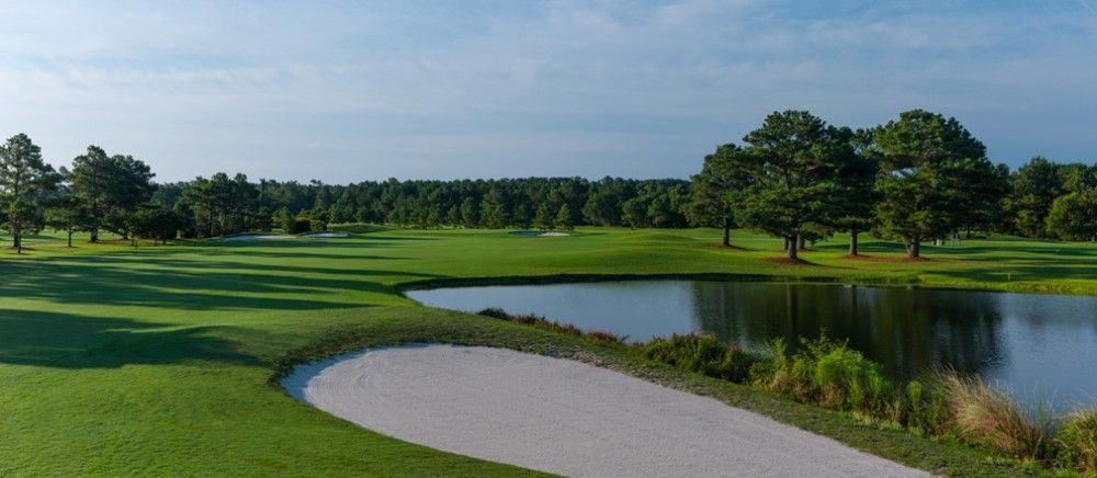Golf course with a sand trap in the foreground, a pond, and green grass leading to trees under a cloudy sky.