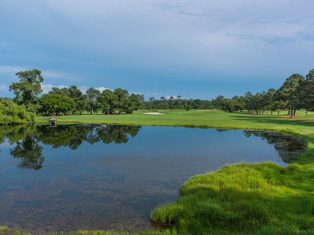 A golf course scene with a reflecting pond in the foreground, green grass, trees, and a cloudy blue sky.