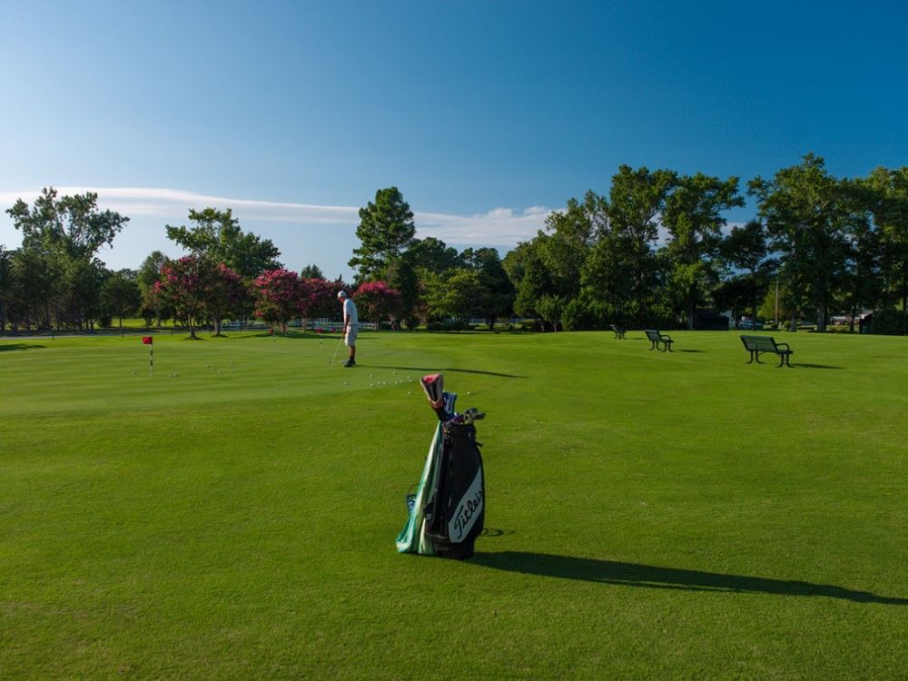 Golf course scene with a golfer putting, golf bag in the foreground, and benches under trees on a sunny day.