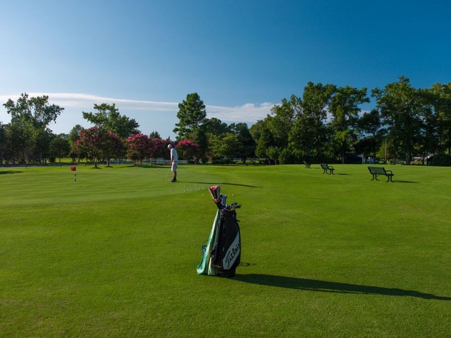 Golf course scene with a golfer putting, golf bag in the foreground, and benches under trees on a sunny day.