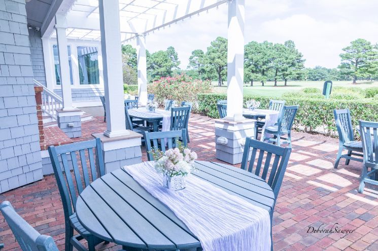Outdoor patio with round tables, blue chairs, and a view of a green lawn. Tables are set for dining, with a floral centerpiece on one.