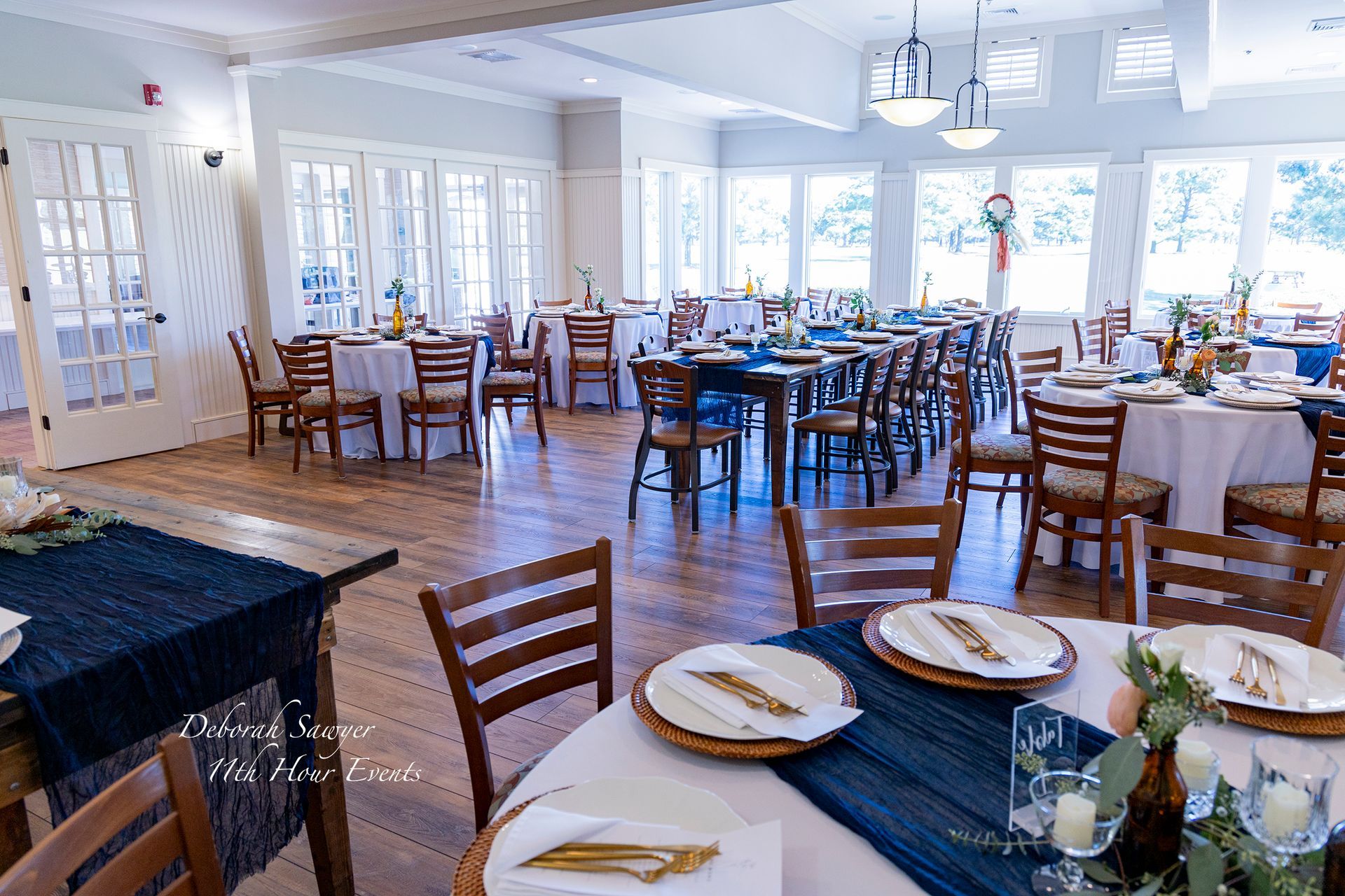 Elegant event space with round and rectangular tables set for dining. Tables are draped in white and blue linens, with chairs and large windows.
