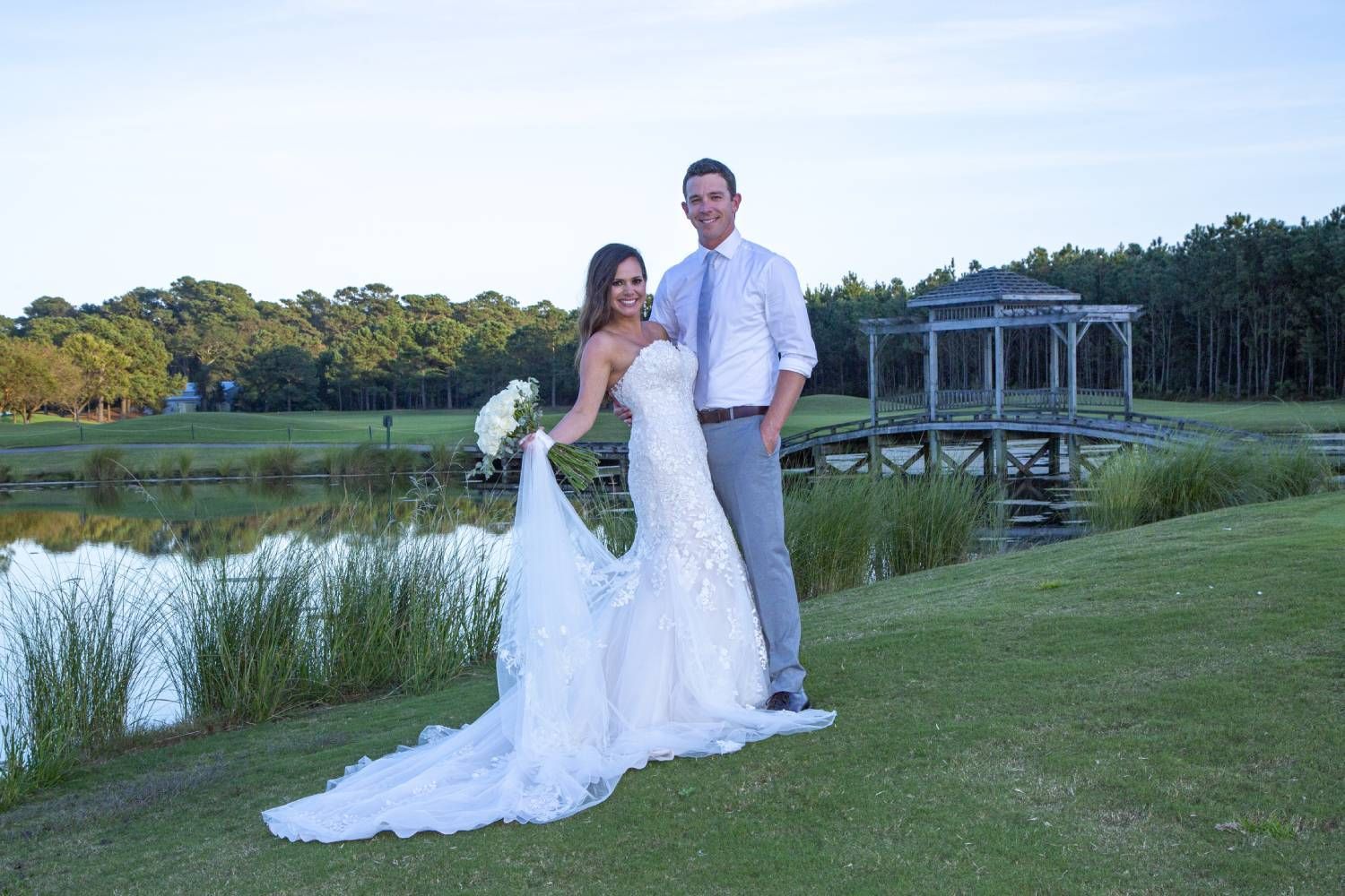 Bride and groom pose by a lake at their wedding. The bride wears a white gown, and the groom wears a light-colored shirt and pants.