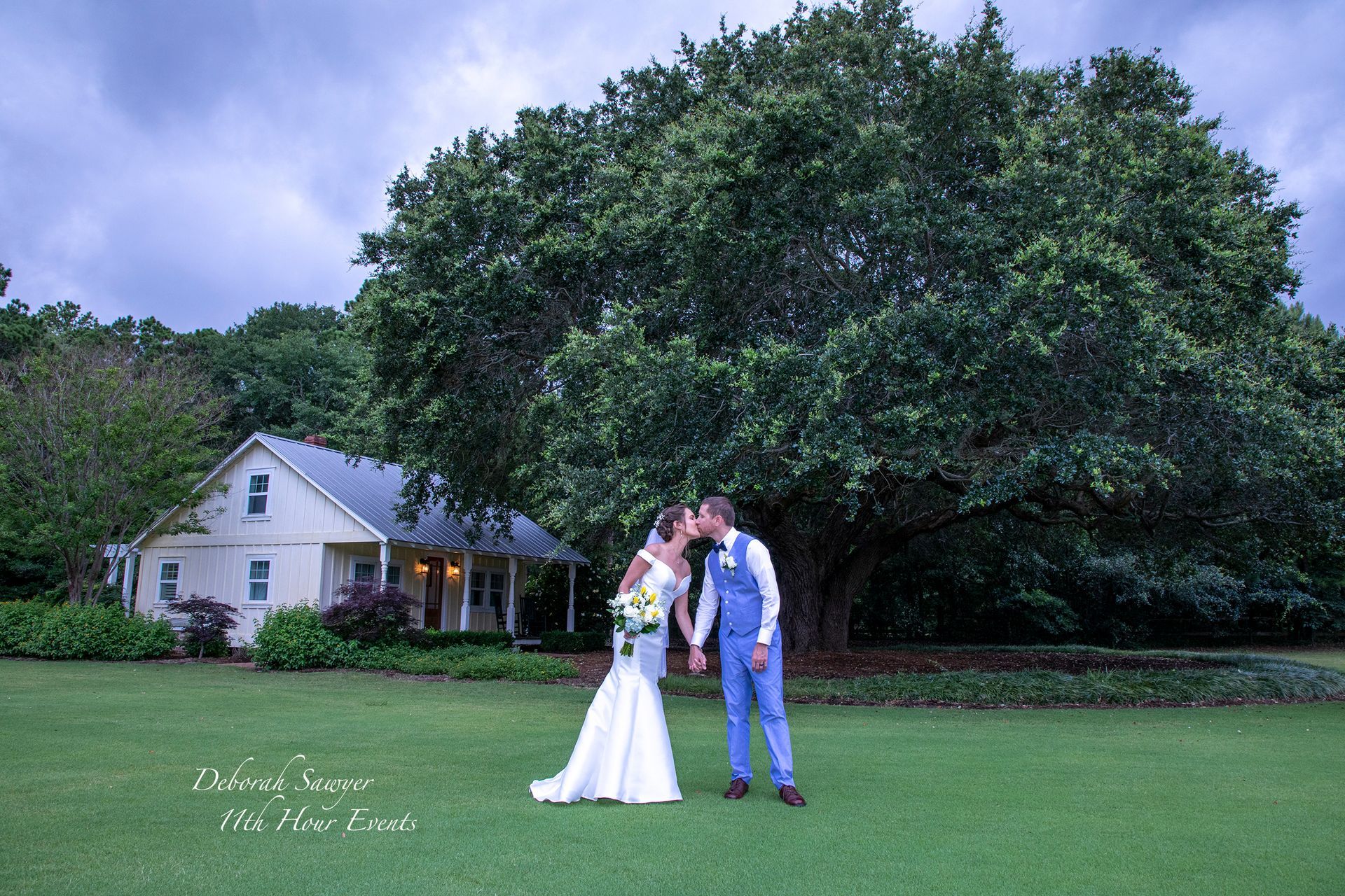 Newlyweds kissing on a green lawn in front of a white house and large tree under an overcast sky.