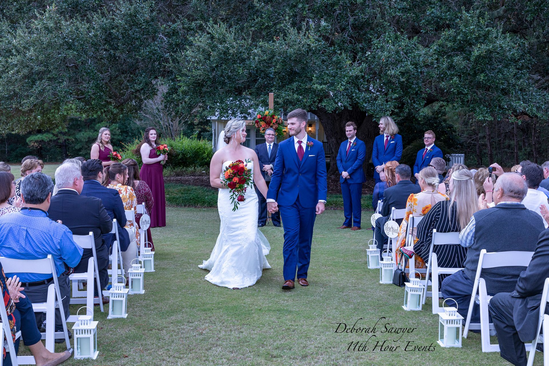 A newlywed couple walks down the aisle after their wedding ceremony. Guests clap, and the wedding party stands behind them.