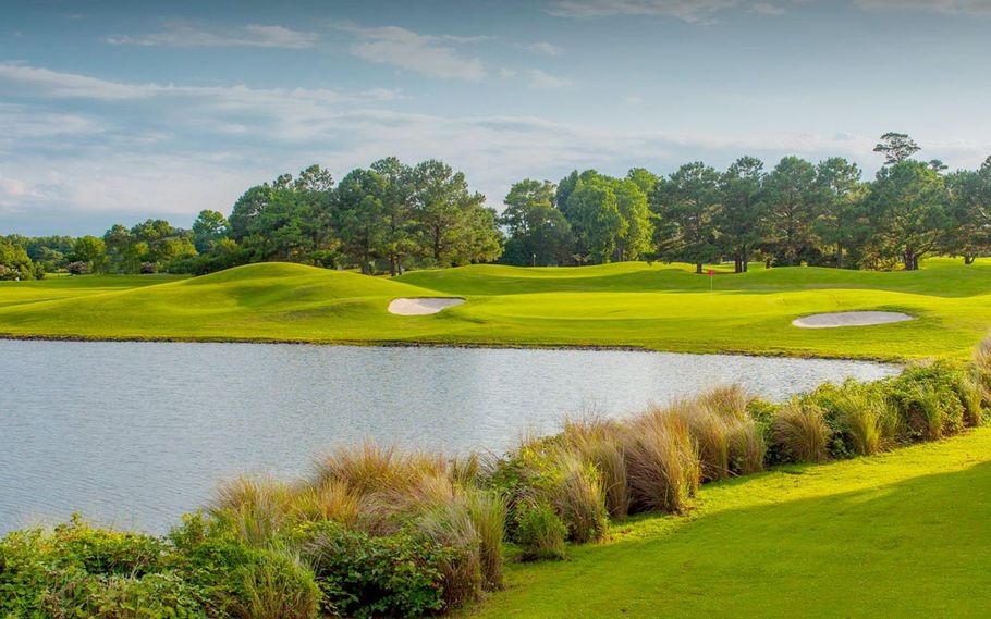 Lush green golf course scene with a pond in the foreground. Trees and a cloudy sky provide a backdrop.