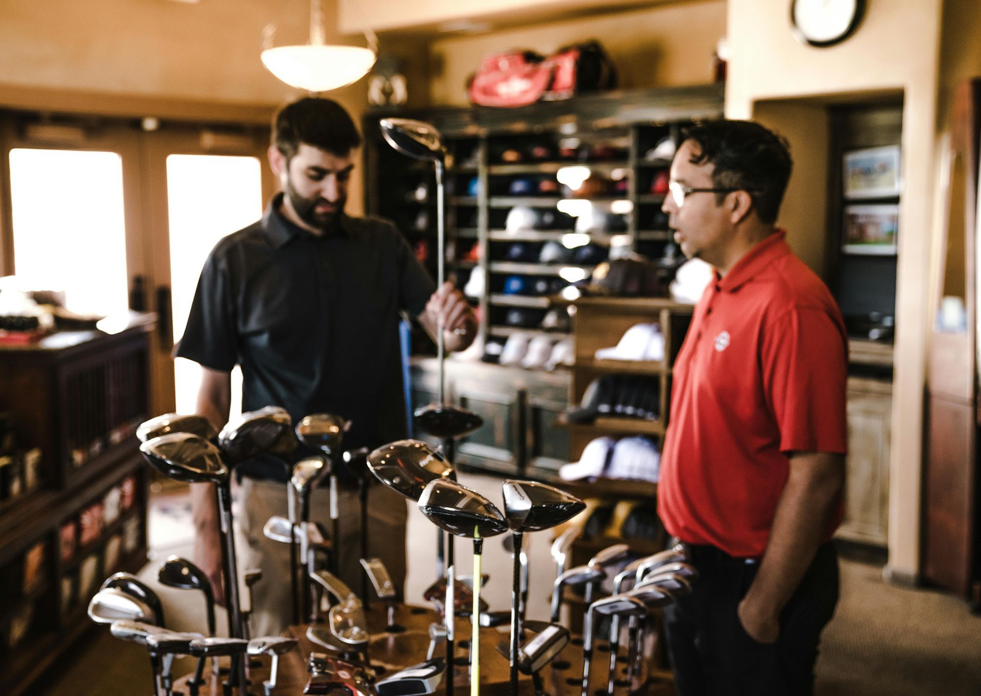 Two men in a golf shop looking at clubs. The shop has shelves of hats and equipment. One man holds up a club.