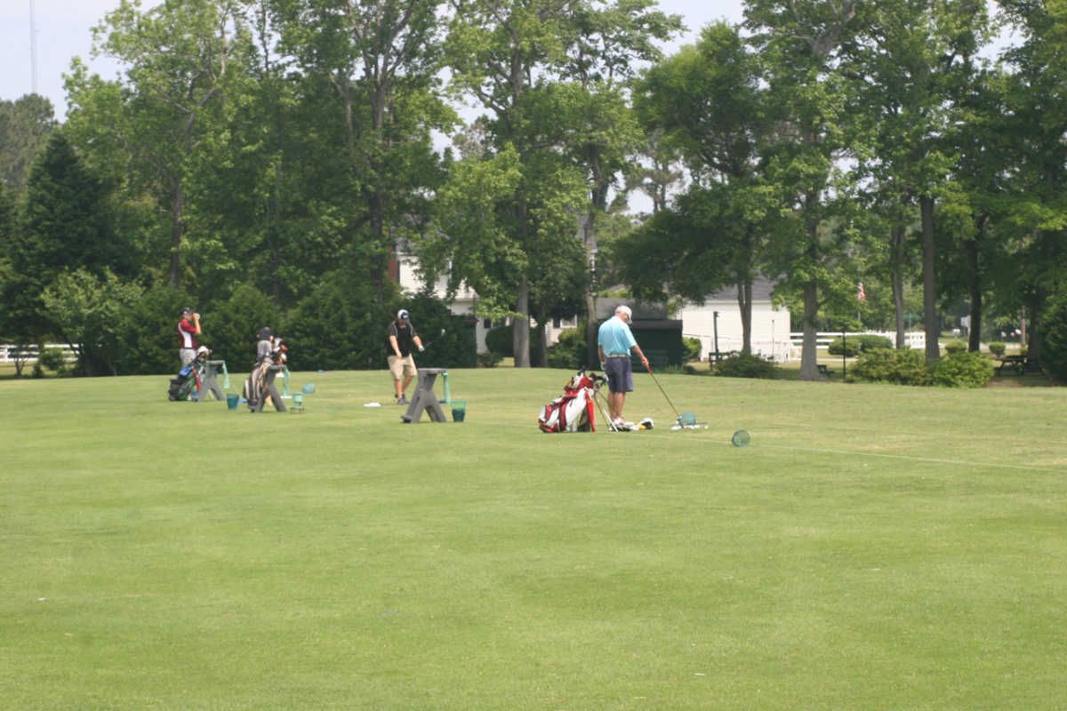 Golfers teeing off on a green lawn with trees in the background. Some golfers are preparing to swing, while others watch.