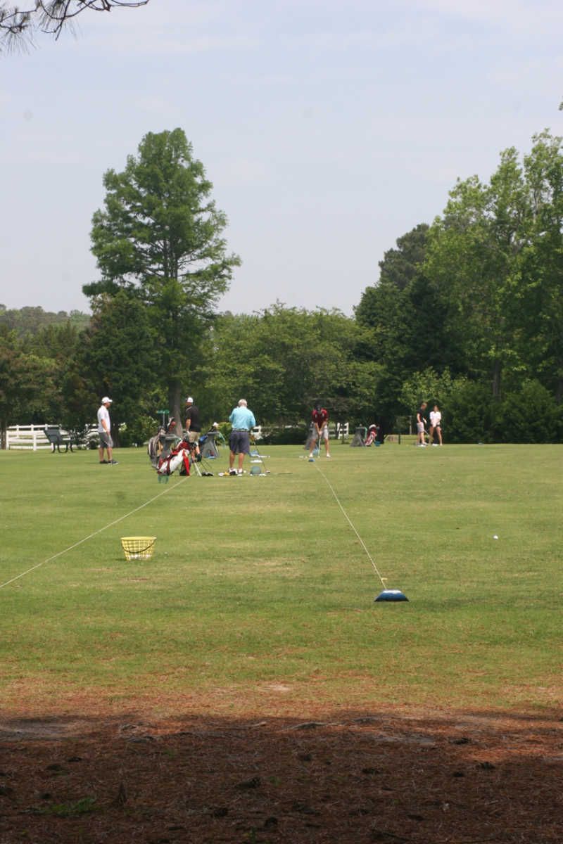 People on a golf course practicing putting. They are in a grassy area with trees in the background, and a blue sky.