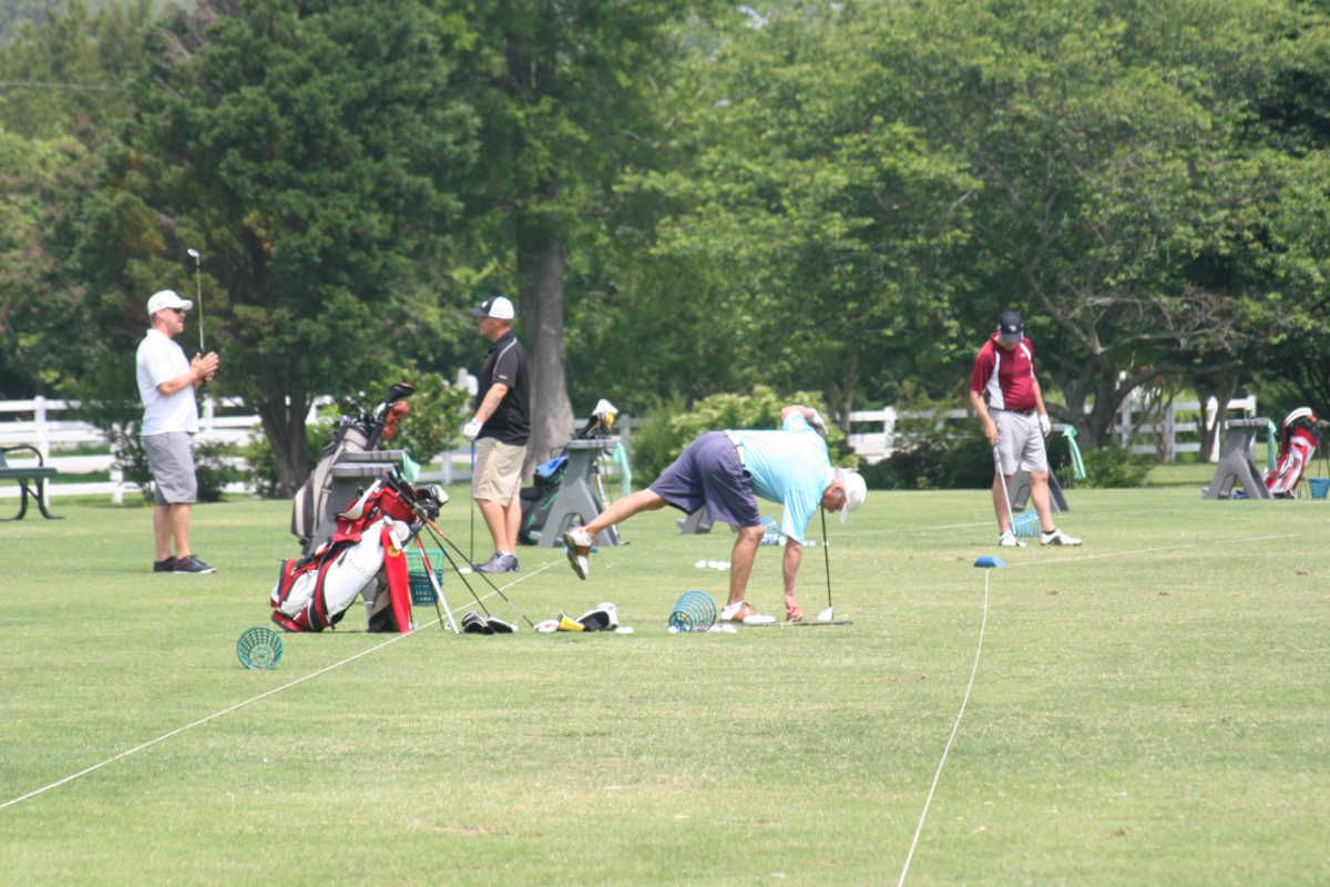 Golfers practicing putting on a green; one is bending over, others watch. Bright, sunny day.