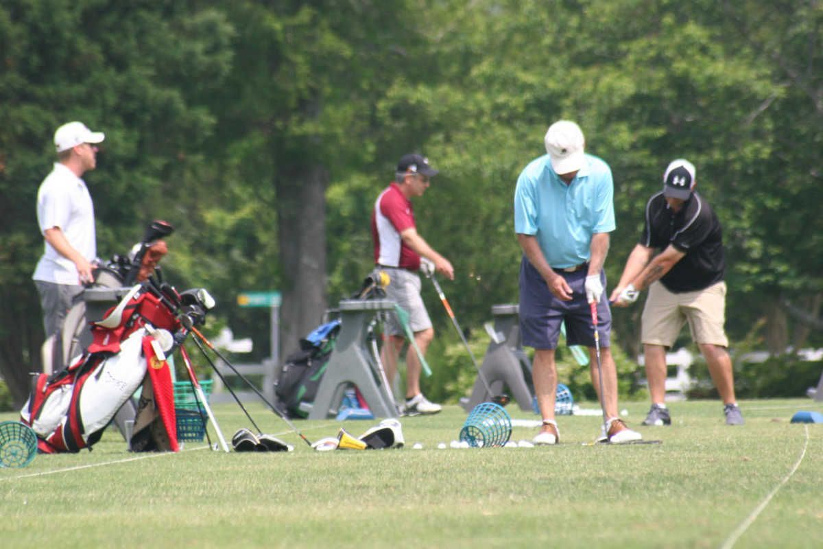 Golfers practicing on a green, sunny day. Men in casual attire prepare to swing clubs, with golf bags and trees in the background.