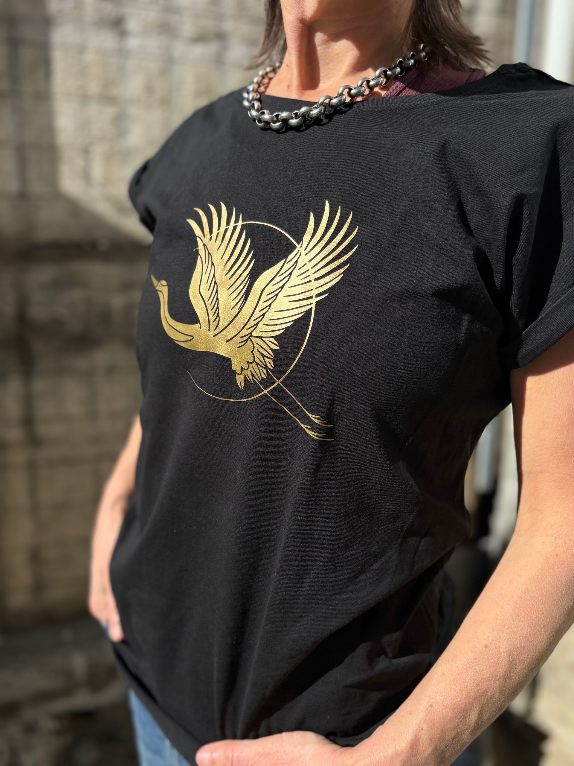 Een persoon draagt een zwart T-shirt met een gouden vogelmotief en een kralenketting, buiten.