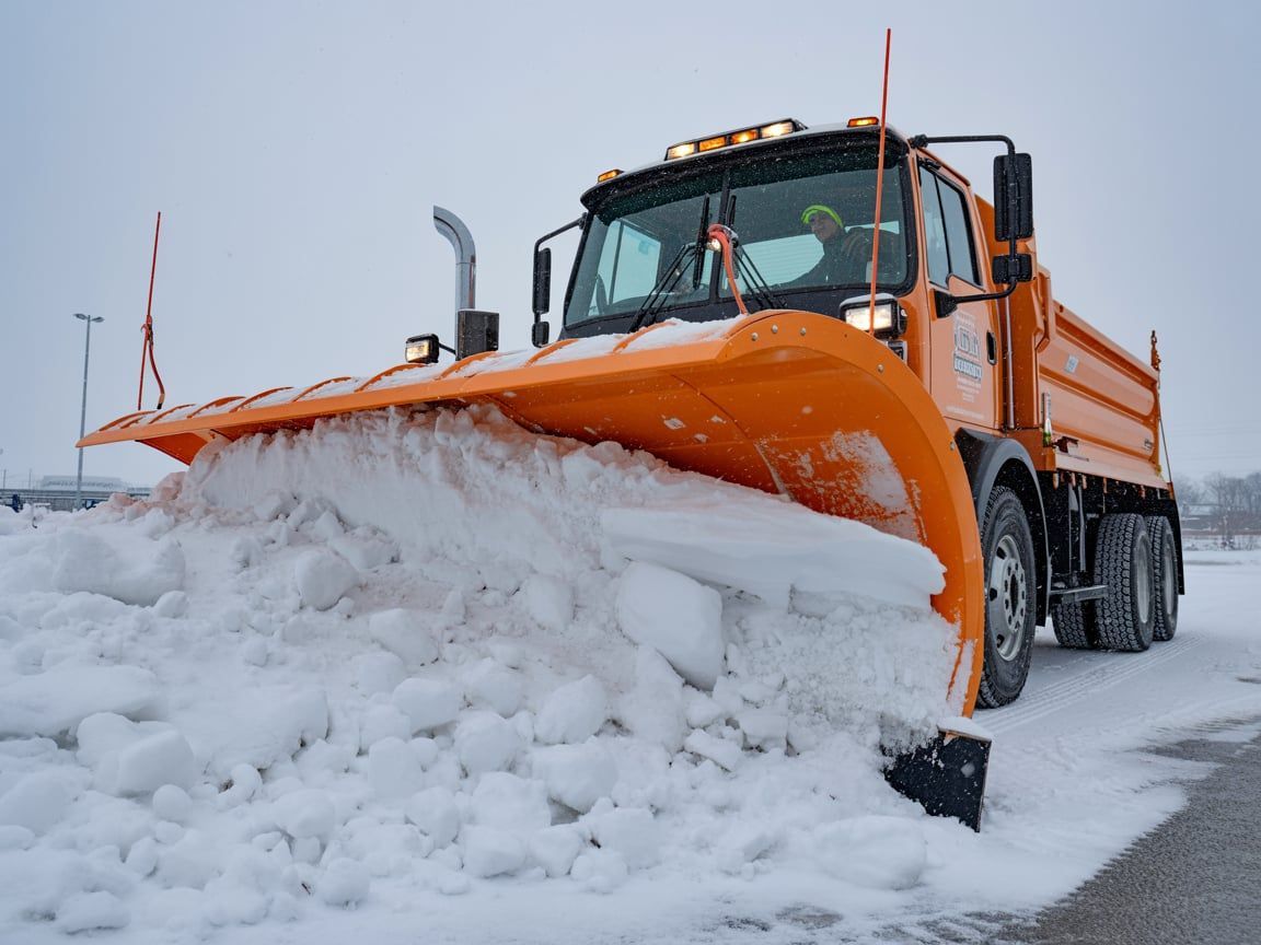 Orange snowplow clearing snow from a parking lot on a gray winter day.