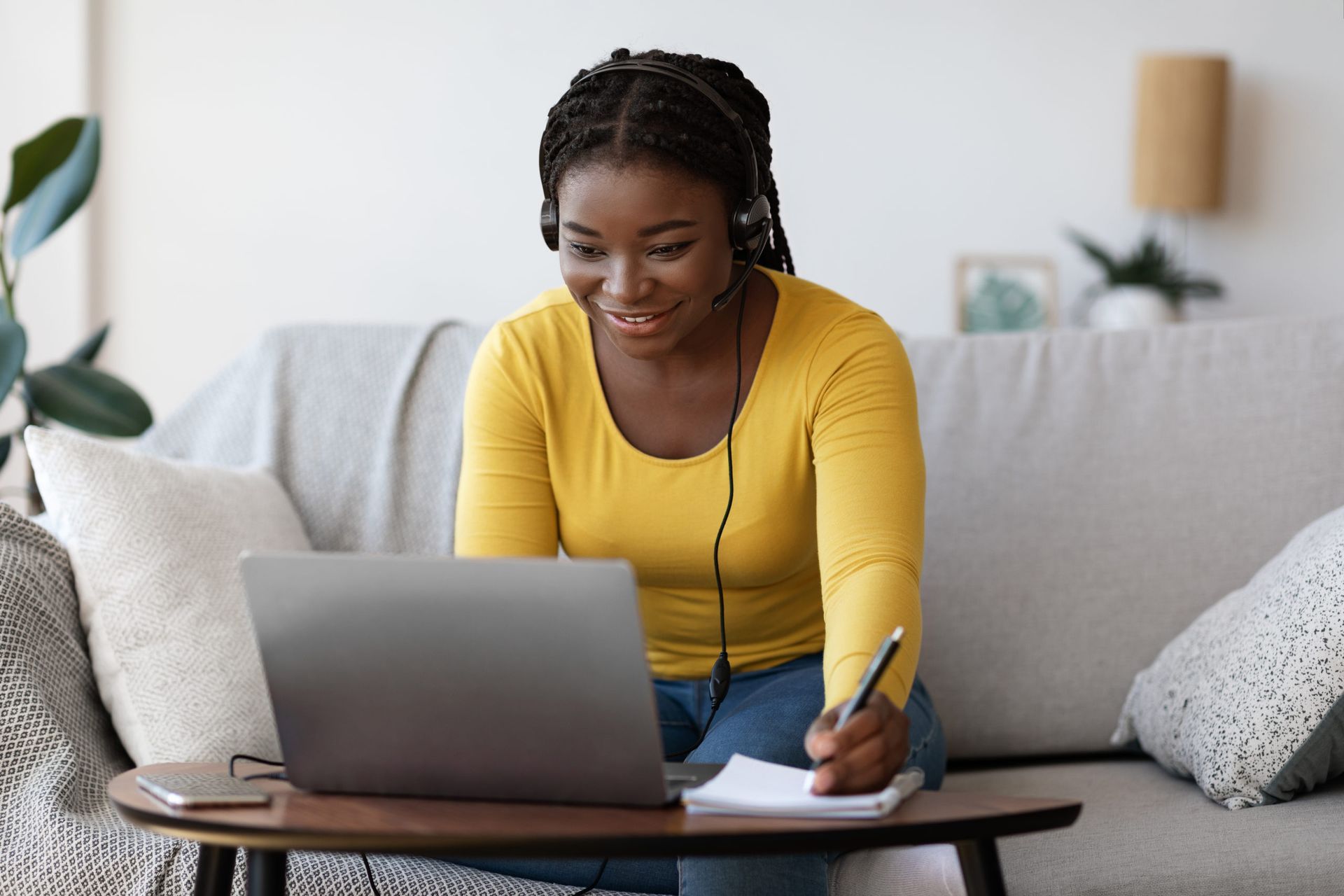 A woman is sitting on a couch using a laptop computer and writing in a notebook.