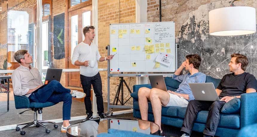 A group of people are sitting on a couch and a man is standing in front of a whiteboard.