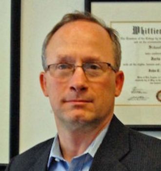 A man wearing glasses and a suit stands in front of a framed diploma